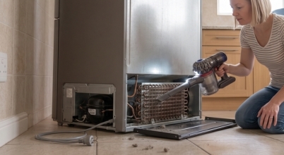 A woman vacuums cleans dust from the condenser coils on an unplugged fridge